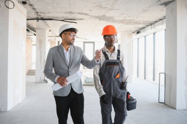 Mimarın elinde bina içinde alet çantası ve telsiz taşıyanlara talimatlar gösteren planlar var.