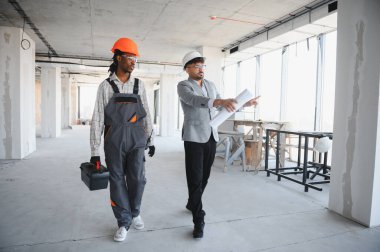 İnşaat alanından geçen bir mimar, gelişmekte olan yapıyı incelerken bir inşaatçıyla planları tartışıyor.