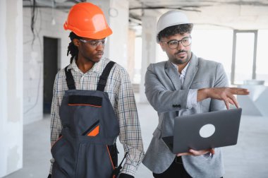 İnşaat teftişi sırasında bir inşaatçıya dizüstü bilgisayardaki bina planlarını sunan mimar, işbirliği ve iletişimi teşvik ediyor.