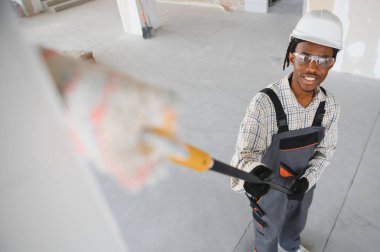 Afrikalı Amerikalı inşaat işçisi duvarları boyuyor ve teleskopik boya silindiri kullanarak işyerinde gülümsüyor.