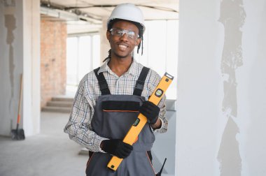 Gülümseyen Afro-Amerikan inşaat işçisi güvenlik kaskı takıyor ve bir inşaat alanında ruh seviyesini koruyan tulumlar giyiyor.