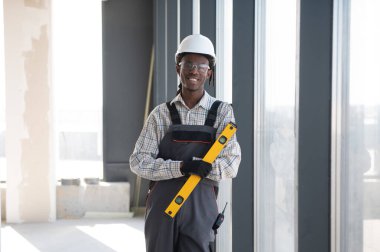 Afrika kökenli Amerikalı inşaat işçisi. Güvenlik kaskı takıyor ve iş tulumları giyiyor. İnşaat alanında gülümseyen bir ruh seviyesinde.