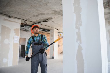 İnşaat işçisi teleskopik boya silindiri kullanıyor, inşaat halindeki bir binanın içine duvar boyuyor.