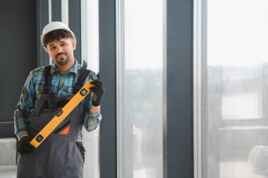 Genç inşaat işçisi üniforma giyiyor ve inşaat halindeyken ruh seviyesini sabit tutuyor.