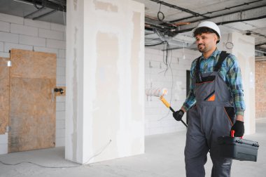 Arap mühendis boya silindiri ve alet çantası taşıyarak inşaat sahasında yürüyor, tulum ve şapka takıyor.