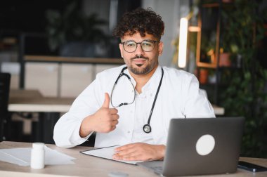 Kendine güvenen Arap erkek doktor, modern klinikteki onay ve memnuniyetini ifade ederek dizüstü bilgisayarla ve tıbbi belgelerle masaya otururken elini kaldırıyor.
