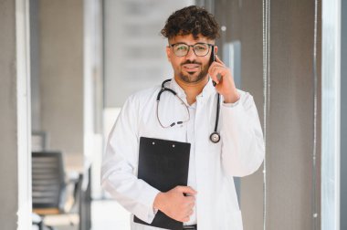 Laboratuvar önlüğü ve steteskop giymiş gülümseyen Arap doktor elinde pano, hastane koridorunda telefonla konuşuyor.