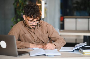 Genç Arap öğrenci yanında dizüstü bilgisayarla üniversite kütüphanesinde okurken deftere yazı yazıyor.