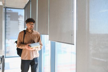 Genç Arap öğrenci üniversite koridorunda yürüyor, kitap ve sırt çantası taşıyor, akademik takip ve büyüme ruhunu somutlaştırıyor.