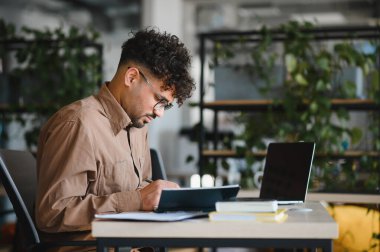 Üniversite kütüphanesinde okurken not yazan genç bir Arap öğrenci, dizüstü bilgisayar ve kitaplarla masada.