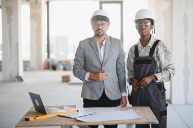 Güvenlik kaskı takan mimar ve müteahhit planları inceliyor ve inşaat alanındaki proje planlarını tartışıyor.