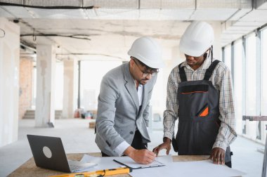 Mimar ve müteahhit başlık takıyor, planları tartışıyor ve hareketli bir inşaat sahasının planları üzerinde işbirliği yapıyor.