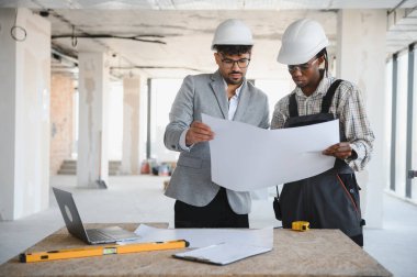 Mimar ve müteahhit, ikisi de şapka takıyor, hareketli bir inşaat alanının ayrıntılı planlarını birlikte inceliyor.