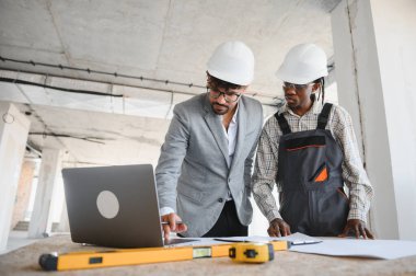 Mimar ve müteahhit başlık takıyor, hareketli bir şantiyede iş birliği yaparken dizüstü bilgisayarın planlarını inceliyor.
