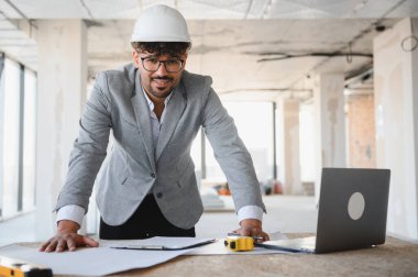 Arap mimar, başlık takıyor, planlara yaslanıyor ve yoğun bir inşaat alanında dizüstü bilgisayarla çalışıyor, proje detaylarını denetliyor.