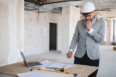 Arap mimar takım elbise ve şapka giyiyor, telsiz kullanıyor, bir inşaat projesini yönetiyor ve planlıyor, planları denetliyor ve inşaat alanında dizüstü bilgisayar kullanıyor.