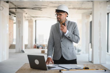 Arap mimar takım elbise ve şapka giyiyor. Dizüstü bilgisayar ve telsiz kullanıyor. İnşaat alanında iletişim kuruyor.