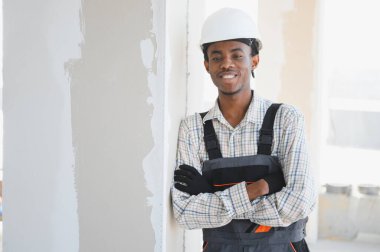 Çapraz kollarla gülümseyen, iş tulumu giyen, inşaat alanında duvara yaslanan genç bir inşaat işçisi.