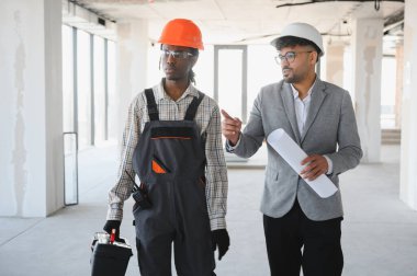 İnşaat halindeki bir binada alet çantası taşıyan inşaatçıya ayrıntıları gösteren planlar tutan mimar