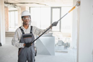 Genç Afro-Amerikan inşaat işçisi inşaat halindeki bir binada teleskopik boya silindiri kullanarak duvarları boyuyor.