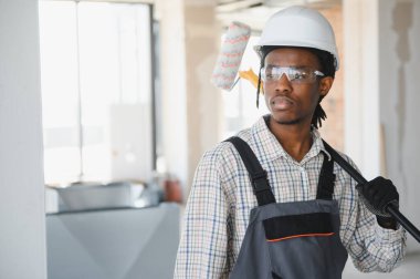 Genç Afro-Amerikan inşaat işçisi omzunda boya silindiri taşıyor, güvenlik gözlüğü ve şapka takıyor, inşaat alanında çalışıyor.