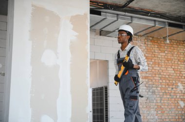 Genç Afrikalı Amerikalı inşaatçı inşaat alanında yürüyor ve elinde üniforma ve şapka takıyor.