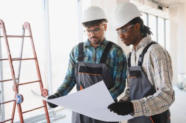 Şapka ve tulum giyen iki mimar, bir planı incelemeye odaklanmış, bir inşaat alanı projesinde işbirliği yapıyorlar.