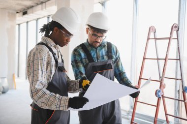 İnşaatta birlikte çalışırken baret ve koruyucu gözlük takan iki müteahhit bina planlarını tutuyorlar.