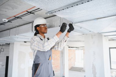 Afrikalı Amerikalı inşaat işçisi inşaat alanına havalandırma boruları yerleştiriyor, tornavida kullanıyor ve güvenlik gözlüğü ve kask takıyor.