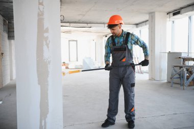 İnşaat işçisi tulum giyiyor ve uzun bir fırça kullanarak duvara alçı yapıştırıyor, inşaat alanında çalışıyor.