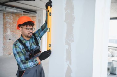 Odaklanmış mühendis, duvarın yüksekliğini ruh seviyesiyle özenle kontrol ederek, hareketli bir inşaat sahasında inşaat standartlarına uyulduğunu ve hassasiyetini gösterir.