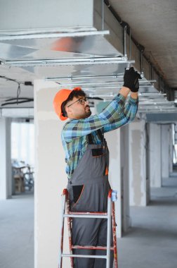 Elektrikçi bir inşaat sahasında havalandırma sistemi kurulumunda çalışıyor, merdiven kullanıyor ve güvenlik ekipmanı takıyor.