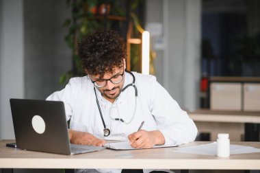 Laboratuvar önlüğü ve steteskop giyen odaklanmış erkek doktor, doktorun ofisinde dizüstü bilgisayar kullanarak reçete yazıyor.