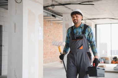 Arap mühendis başlık takıyor, boya silindiri tutuyor ve inşaat alanında yürüyor.