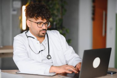 Laboratuvar önlüğü ve steteskop giyen odaklanmış erkek doktor laptopunu kullanıyor, modern klinikte tıbbi kayıtları yazıyor.