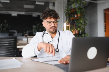 Laboratuvar önlüğü ve steteskop giyen odaklanmış Arap erkek doktor laptop kullanıyor, modern klinikte tıbbi uzmanlık üzerinde çalışıyor.