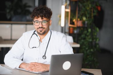 Masasında oturan, sırıtan, steteskopu boynunda ve önünde çalışmaya hazır bir dizüstü bilgisayarla kendinden emin bir Arap erkek doktorun portresi.