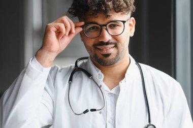Kendine güvenen bir Arap erkek doktorun portresi. Gözlüğünü düzeltiyor, gülümsüyor, beyaz önlük giyiyor ve steteskop takıyor.