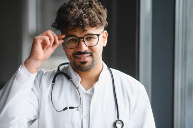 Modern hastane ortamında gözlüklü ve steteskop gülüşlü kendine güvenen Arap erkek doktor.