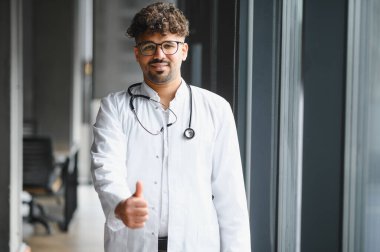 Gülümseyen Arap erkek doktor, modern hastane koridorunda güvenle dikilirken, pozitiflik ve profesyonellik saçarak başparmağını kaldırıyor.