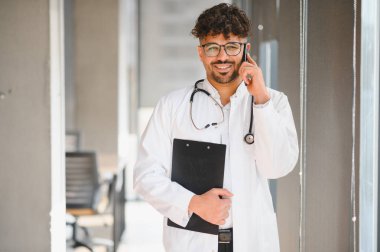 Beyaz önlüklü ve steteskoplu gülümseyen Arap doktor akıllı bir telefonla konuşuyor ve modern bir hastane koridorunda elinde bir pano tutuyor.