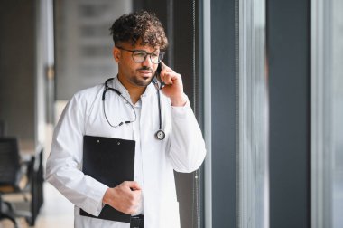 Beyaz önlüklü ve elinde bir pano olan Arap erkek doktor, hastanenin penceresinin yanında bir telefon görüşmesi yapıyor.