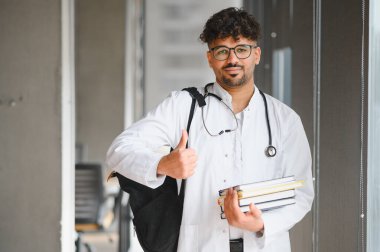 Laboratuvar önlüğü ve steteskop giyen genç ortadoğu tıp öğrencisi üniversite koridorunda el kol hareketi yapan kitaplar ve sırt çantası taşıyor.