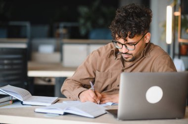 Üniversite kütüphanesinde dizüstü bilgisayarla ders çalışırken notlar yazan genç Arap öğrenci, akademik başarıya odaklandı