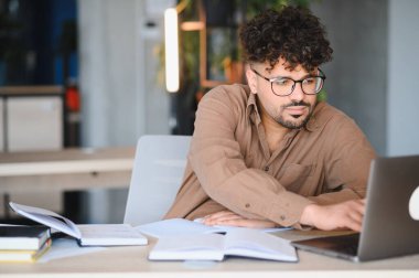 Genç Arap öğrenci laptopuna odaklanmış, parlak bir üniversite kütüphanesinde kitaplarla çevrili, özenle onun çalışmaları üzerinde çalışıyor.