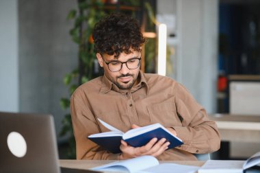 Parlak bir üniversite kütüphanesinde sınavlara hazırlanırken ders kitabı okuyan genç Arap öğrenci, akademik başarı ve bilgiye odaklandı