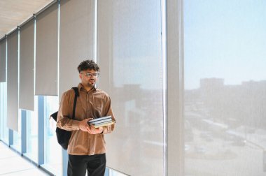 Üniversite koridorunda büyük pencereler ve güneş ışığıyla yürüyen genç erkek öğrenci, öğrenim, eğitim ve okula geri dönüş kavramı