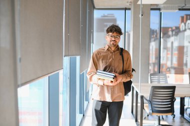 Genç Arap öğrenci bir üniversite koridorunda yürüyor, kitap ve sırt çantası taşıyor, eğitim ve hırs ruhunu somutlaştırıyor