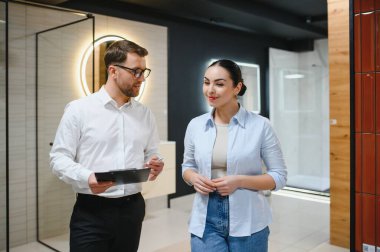 Satıcının elinde bir pano var. Seramik fayans ve su tesisatı dükkanındaki kadın müşteriye banyo malzemelerini gösteriyor.