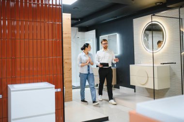 Parlak, modern bir seramik ve tesisat mağazasında müşteriye banyo armatürlerini sunan satıcı seçenekleri ve stilleri tartışıyor.